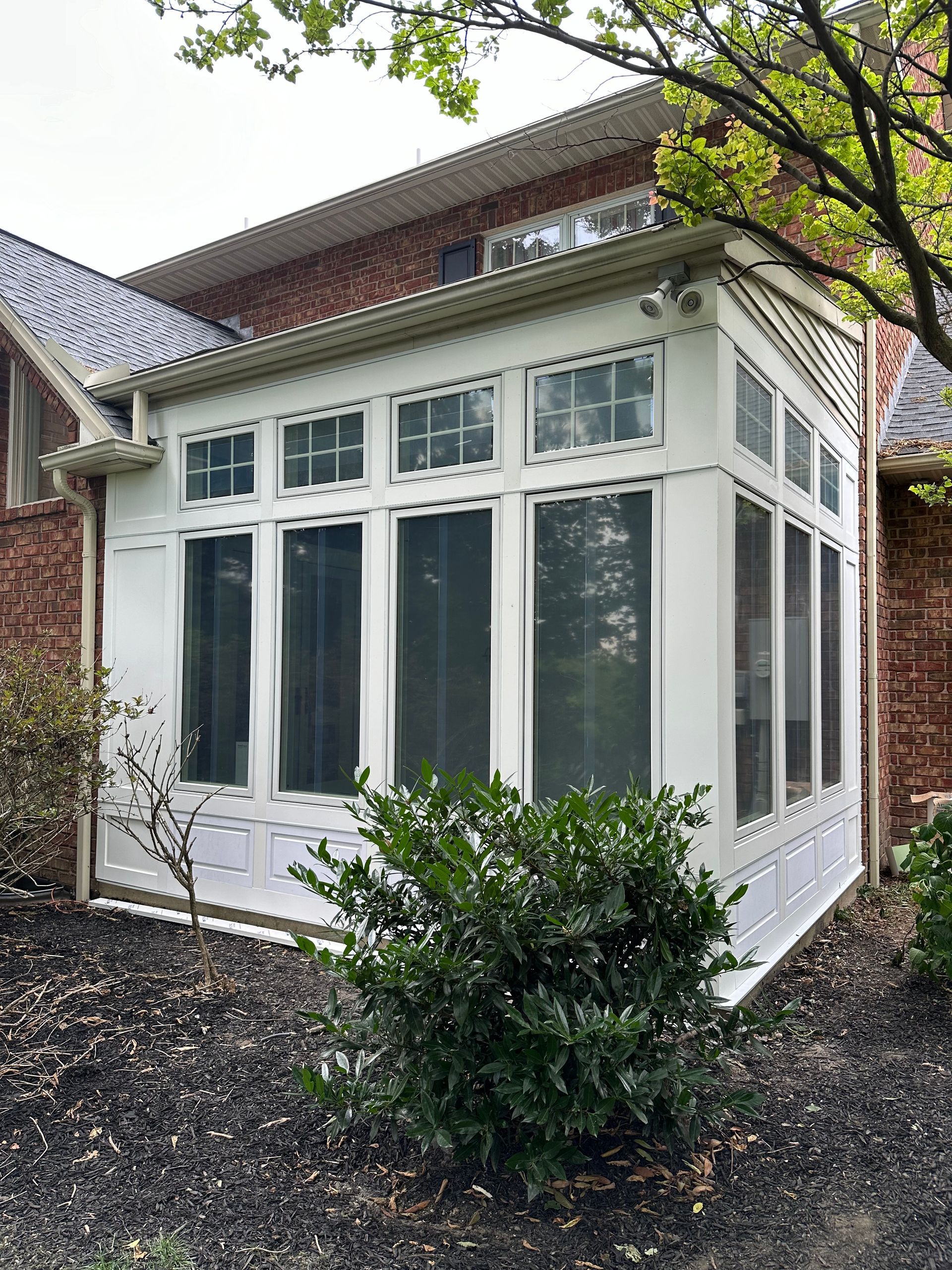 White sunroom addition with large windows attached to a brick house, small bush in the foreground.