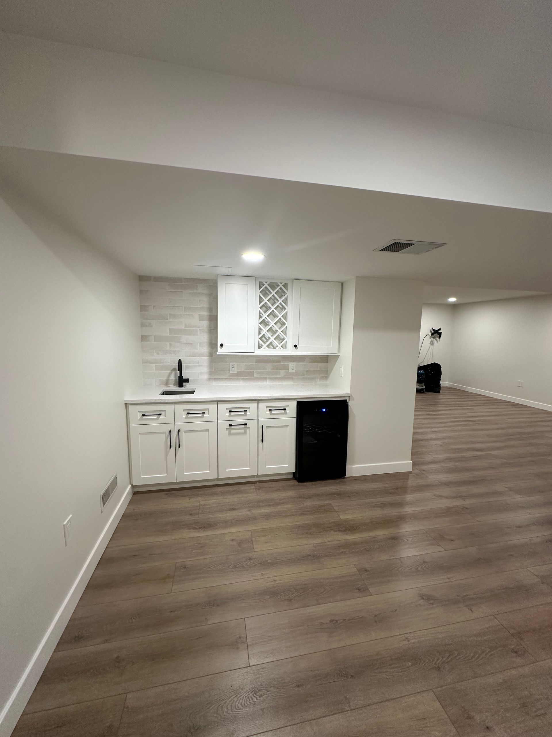White cabinets and backsplash in a basement bar area with flooring and wine cooler.