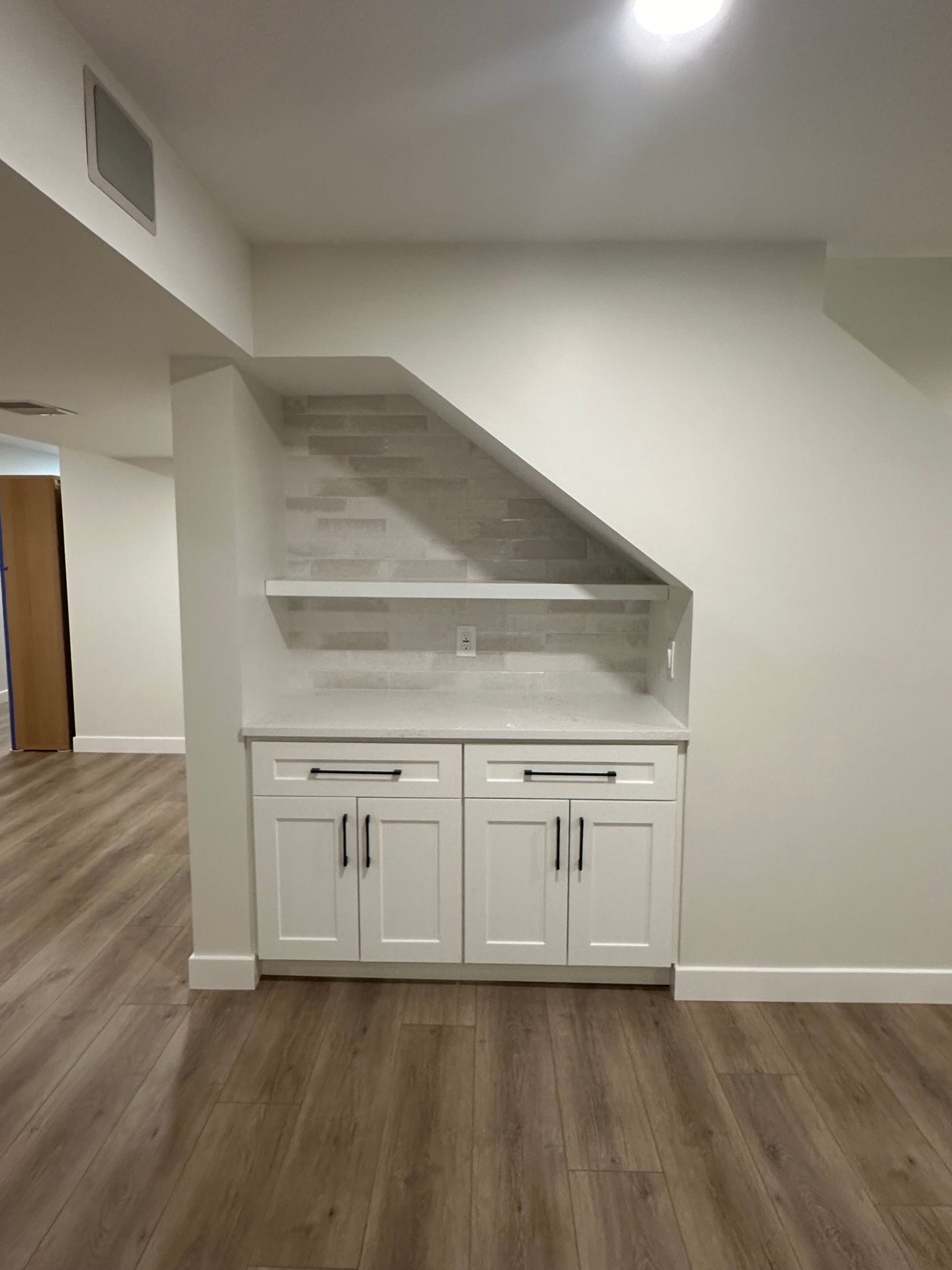 Built-in white cabinets with countertop under a sloped wall with tile, in a room with wood flooring.