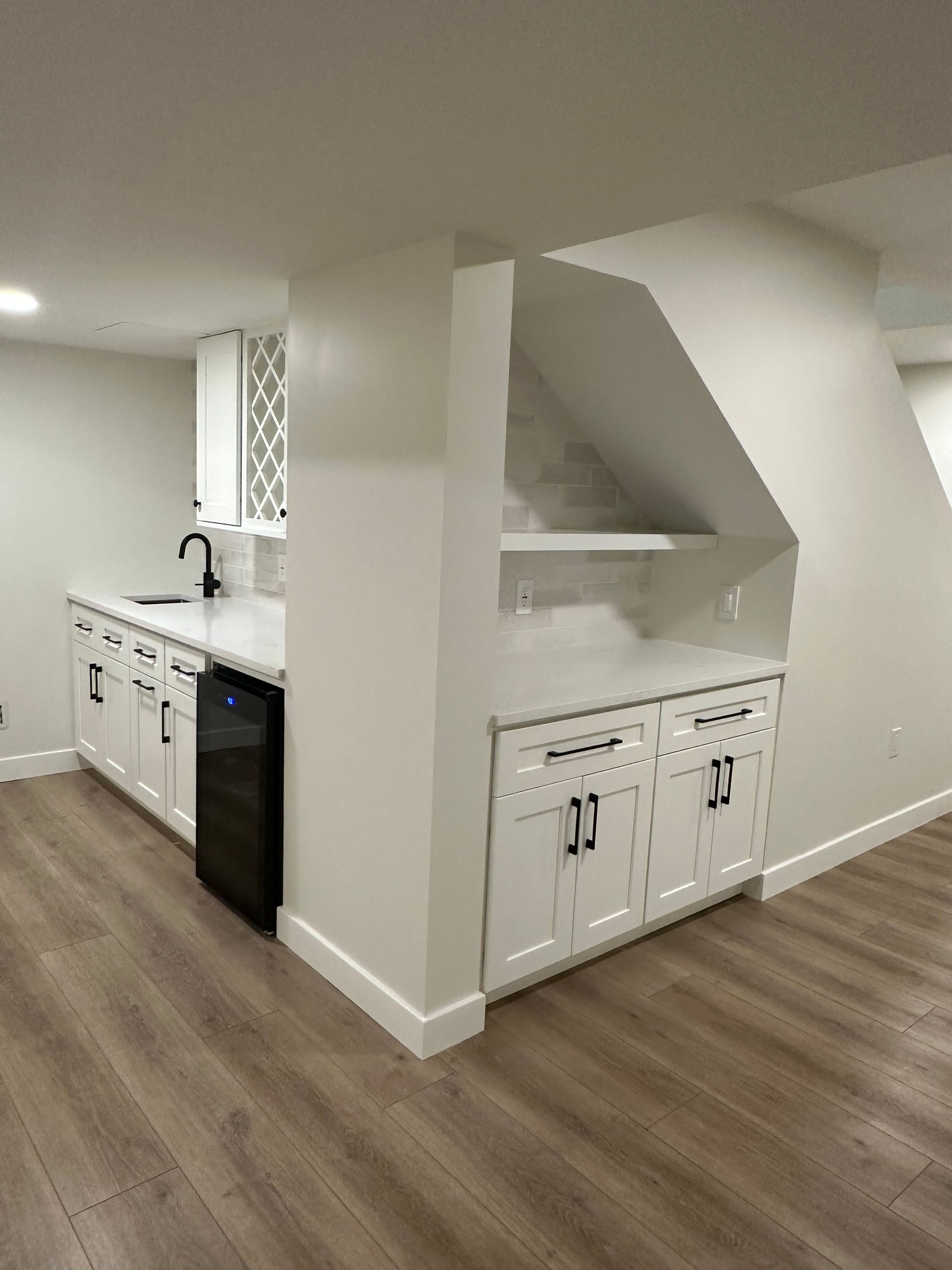 White cabinetry in a basement kitchenette. Black hardware, wine fridge, sink, counter space, and decorative shelf.
