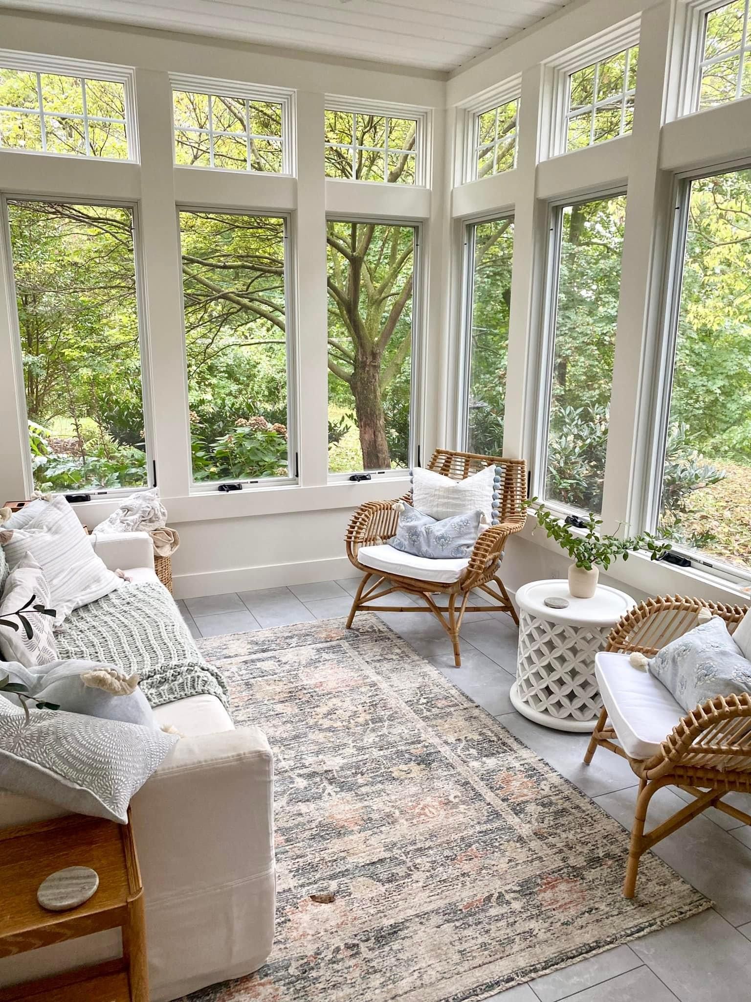 Sunroom with wicker furniture, rug, white walls, and large windows overlooking trees.