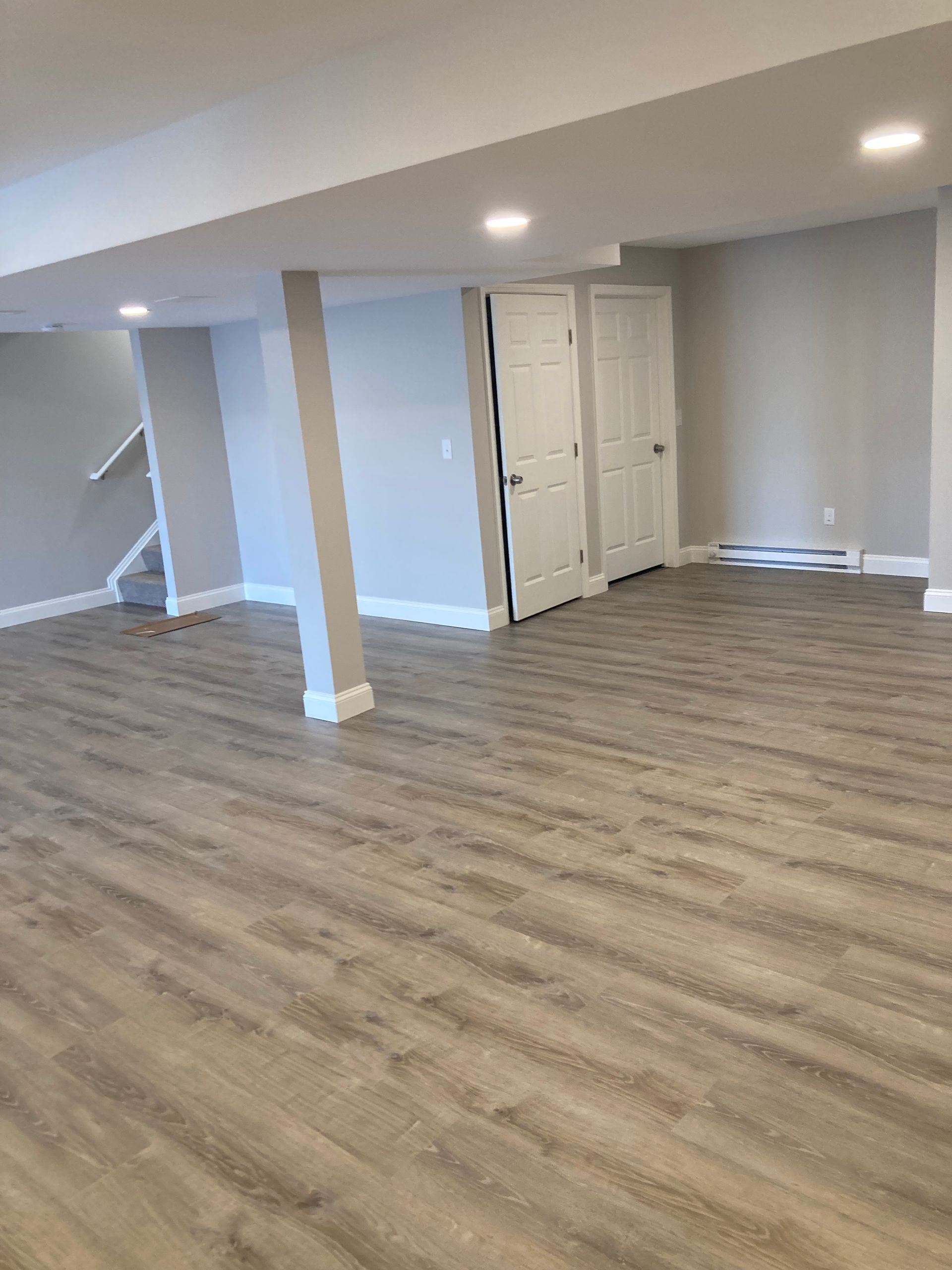 Empty, newly renovated basement with light gray walls, wooden flooring, and two white doors.