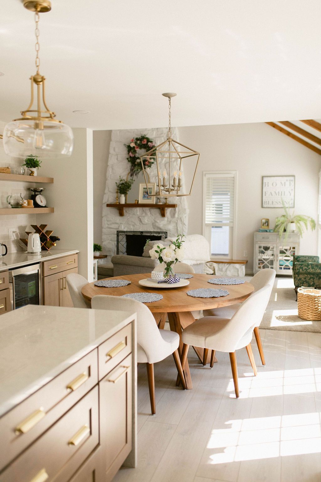 Sunlit dining room with round wooden table, white chairs, and elegant light fixtures.