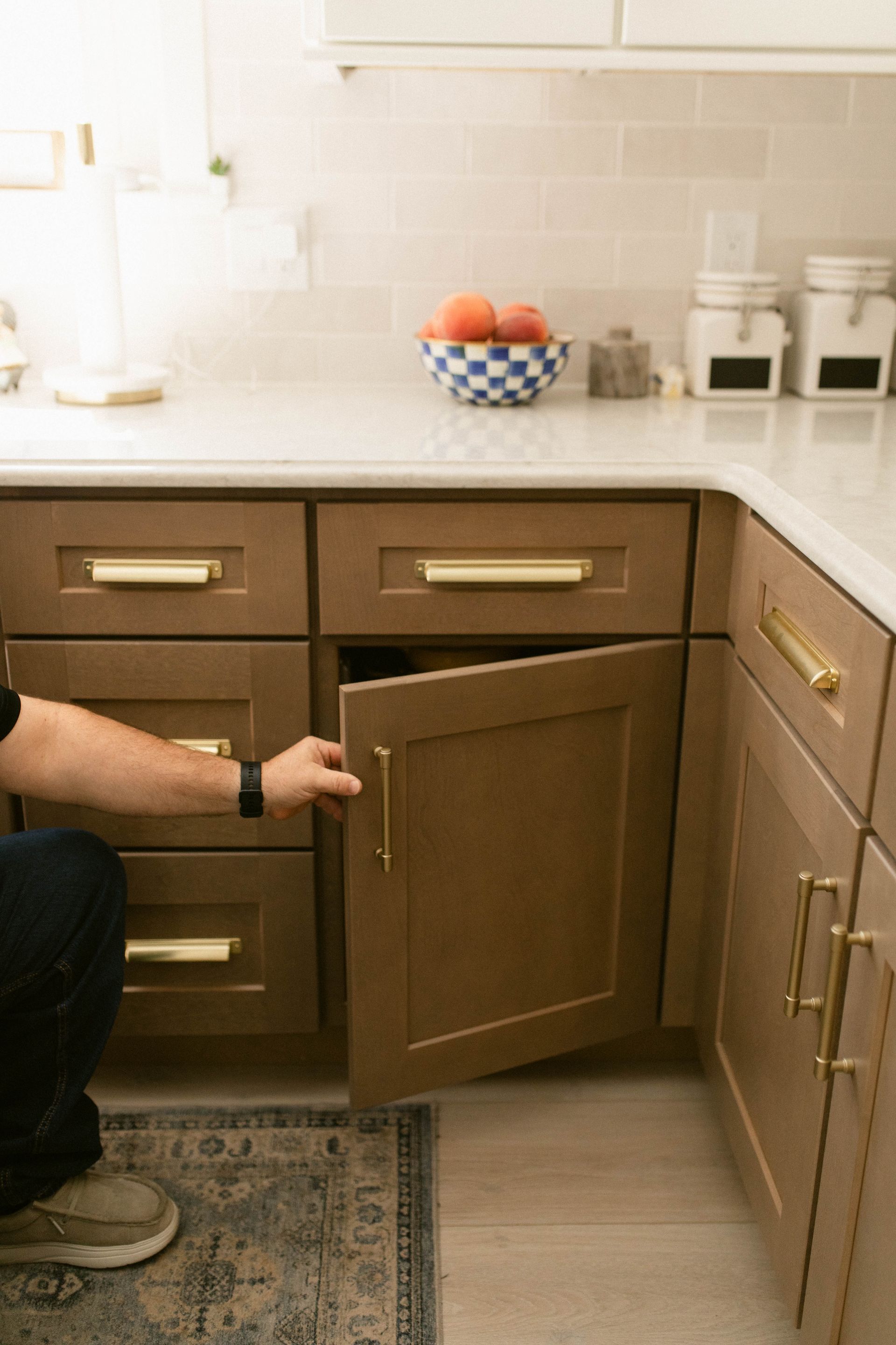 Person opening a brown kitchen cabinet, gold hardware. Kitchen with countertop, fruit bowl, and rug visible.