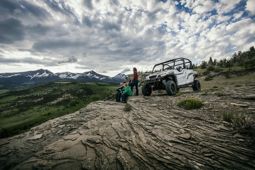 Family admiring mountain view from rocky outcrop, white side-by-side vehicle nearby. Cloudy sky.