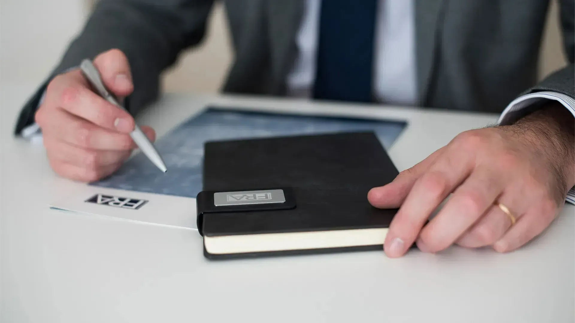 A professional in a suit holds a pen over a document next to a black notebook on a white desk.