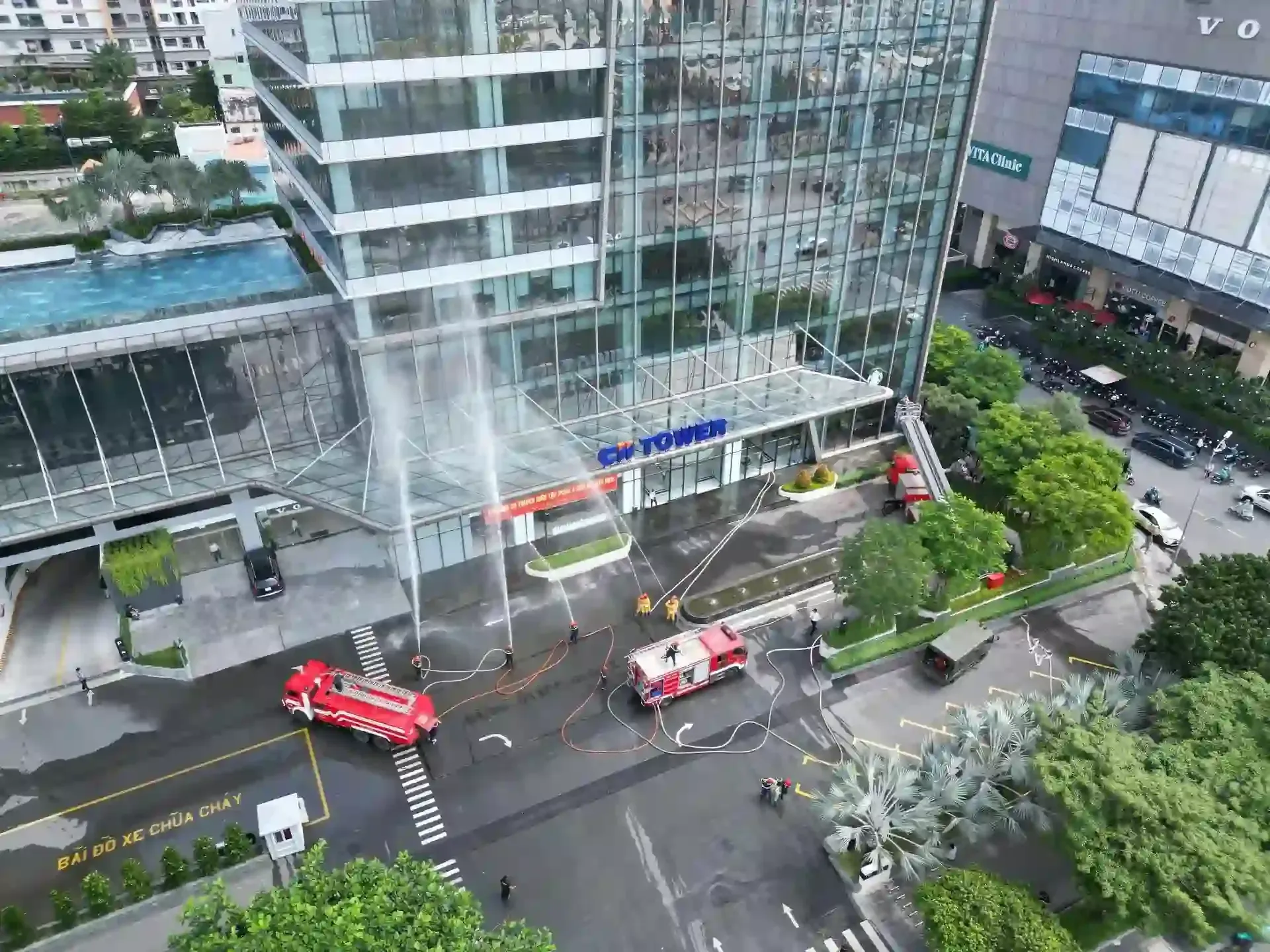 Two red fire trucks spray water onto the glass facade of a city tower while personnel stand nearby in the street.