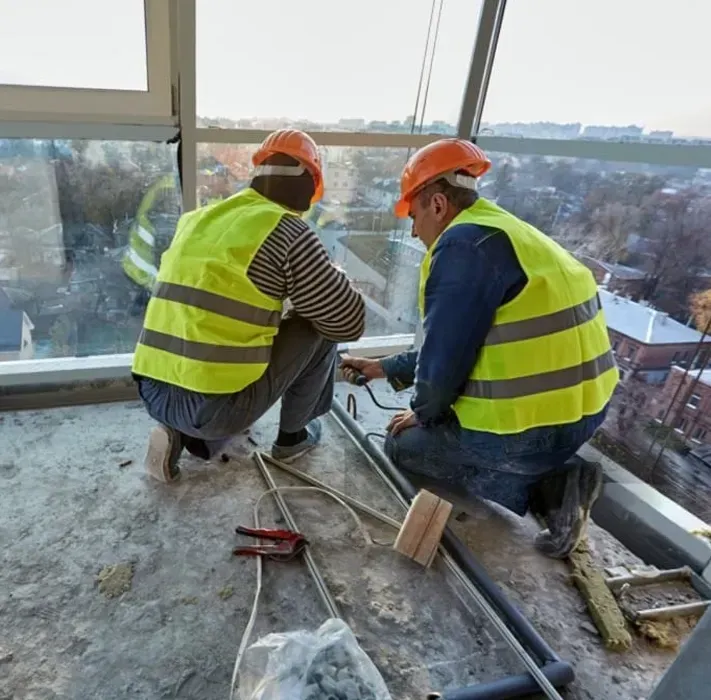 Two construction workers wearing high-visibility vests and hard hats work on an unfinished high-rise building balcony.