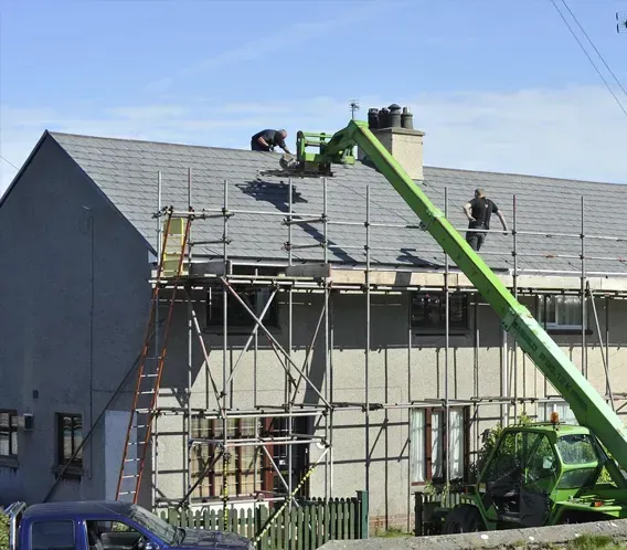 Workers use a bright green telehandler crane to assist with roofing repairs on a house framed by metal scaffolding.