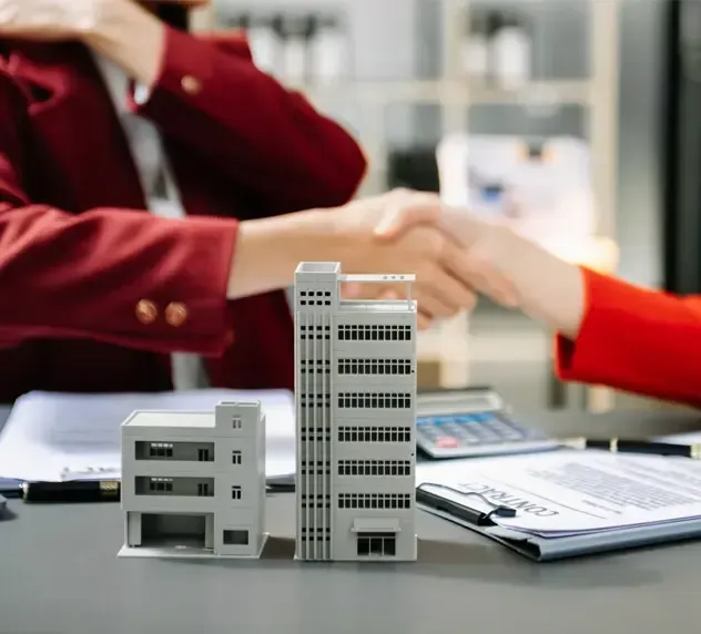 Two people in professional attire shake hands over a desk featuring miniature building models and contract documents.