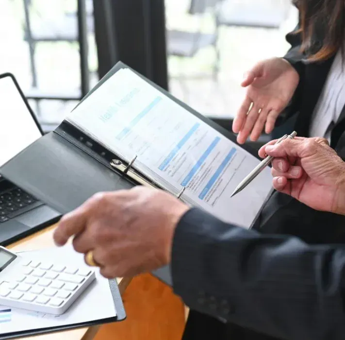 Two professionals in business attire review a document in a binder, with one pointing at the page with a pen.