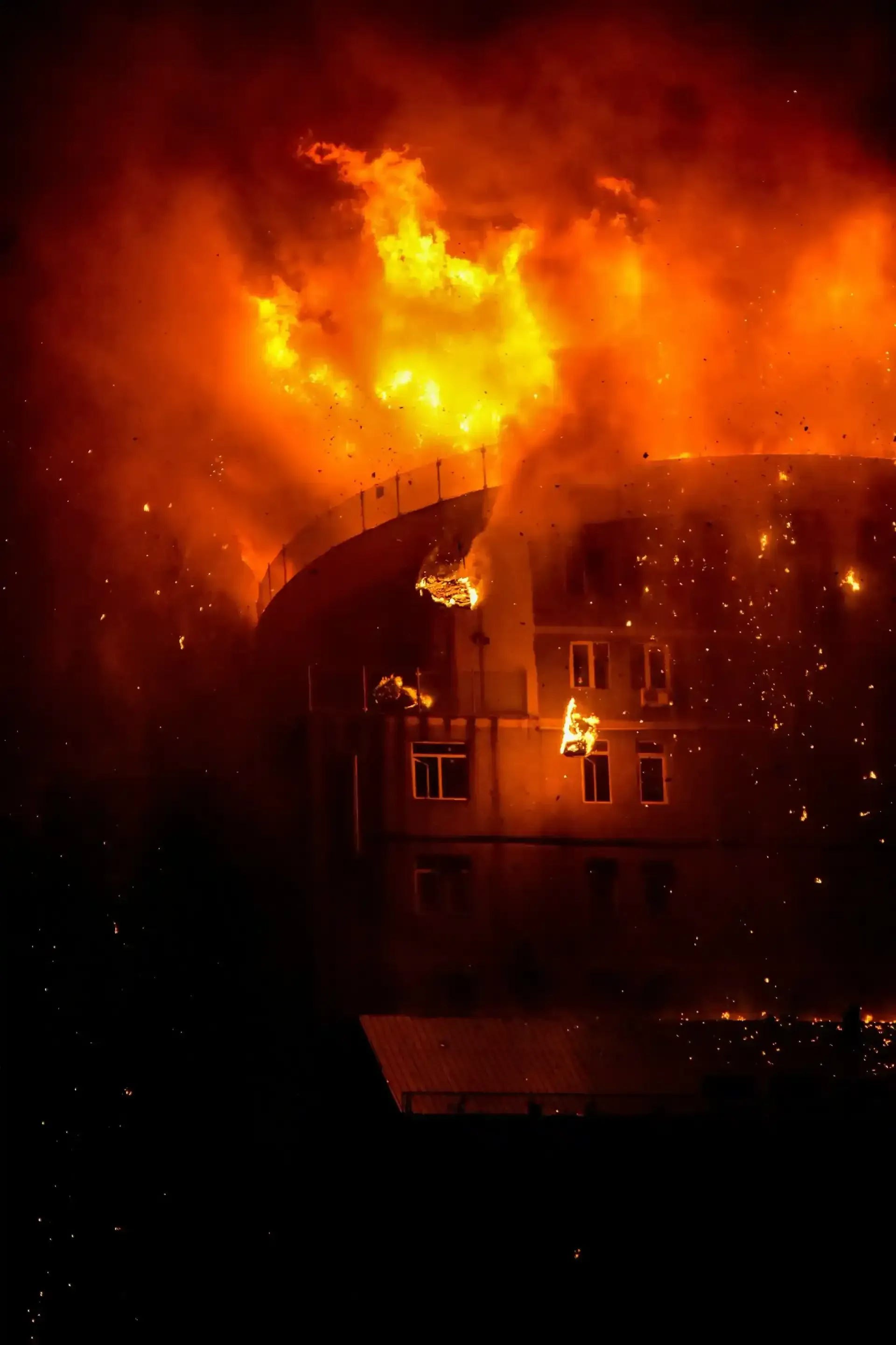 A multi-story building engulfed in intense, bright orange flames during the night, with glowing embers falling.