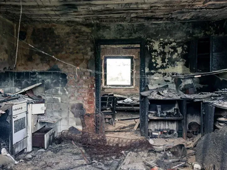 A scorched, debris-filled room with a damaged stove, soot-covered walls, and a doorway leading to a windowed room beyond.