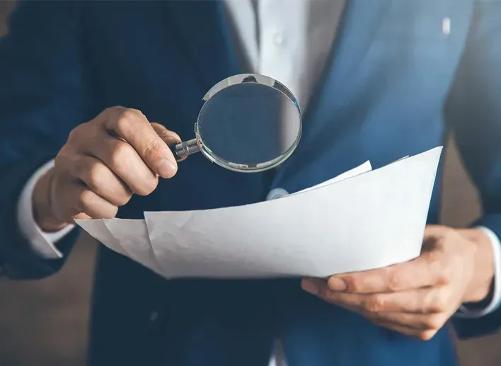 A person in a blue suit uses a magnifying glass to closely examine a stack of white papers.