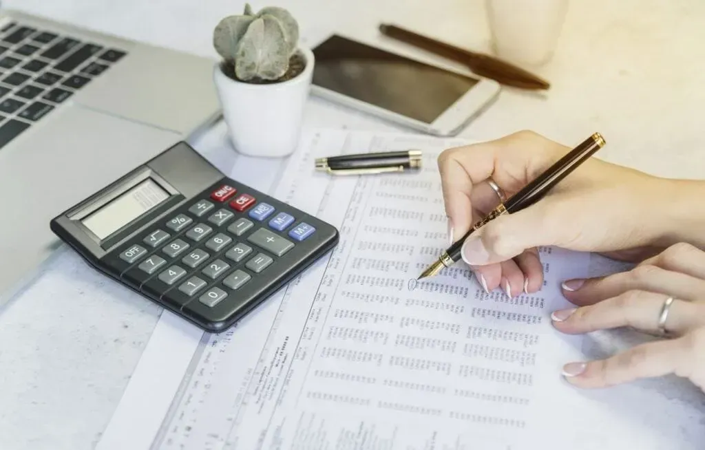 A person writing on a document with a pen next to a calculator, a smartphone, and a small potted plant on a desk.