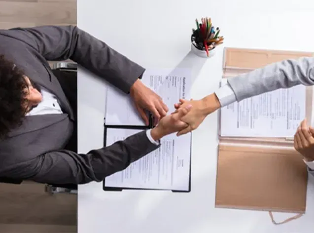 Two professionals in suits shaking hands over a desk with documents and a pen holder, viewed from an overhead angle.