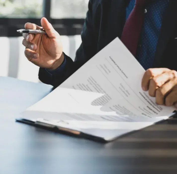 A person in a suit and tie holds a pen while reviewing a document on a clipboard at a desk.