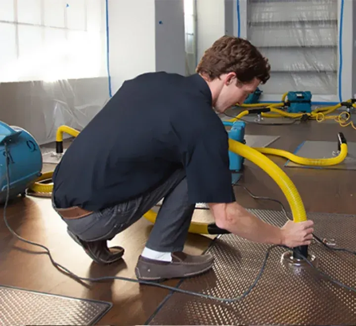 A technician kneels on a floor, connecting a yellow hose to a panel to perform water damage restoration.