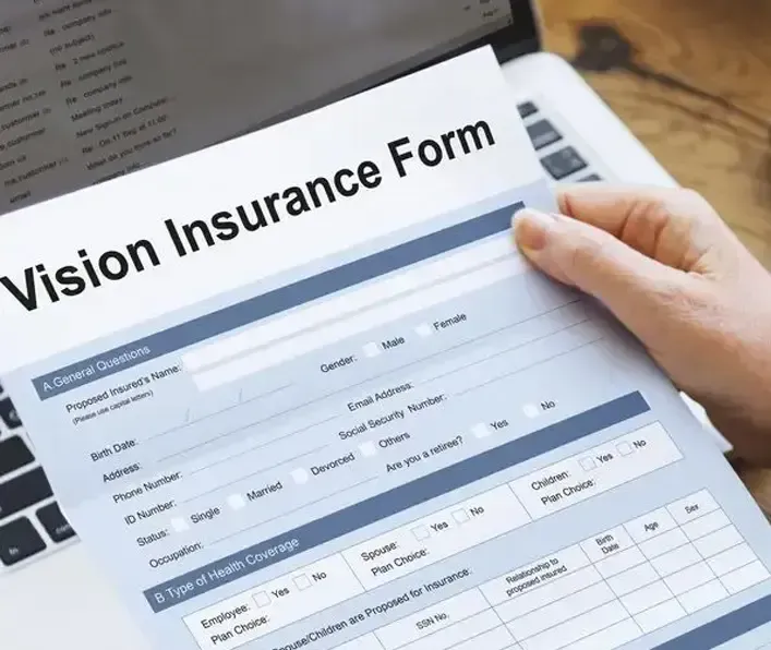 A person's hand holds a paper Vision Insurance Form in front of a laptop computer.