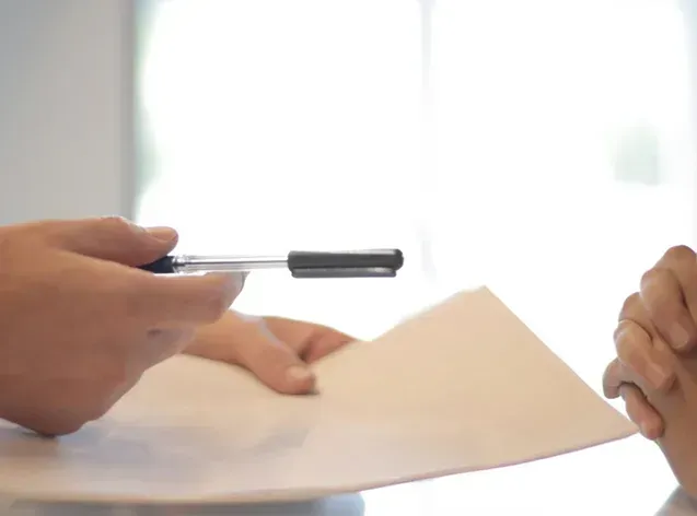 A person holds a pen and points toward a blank sheet of paper during a meeting.