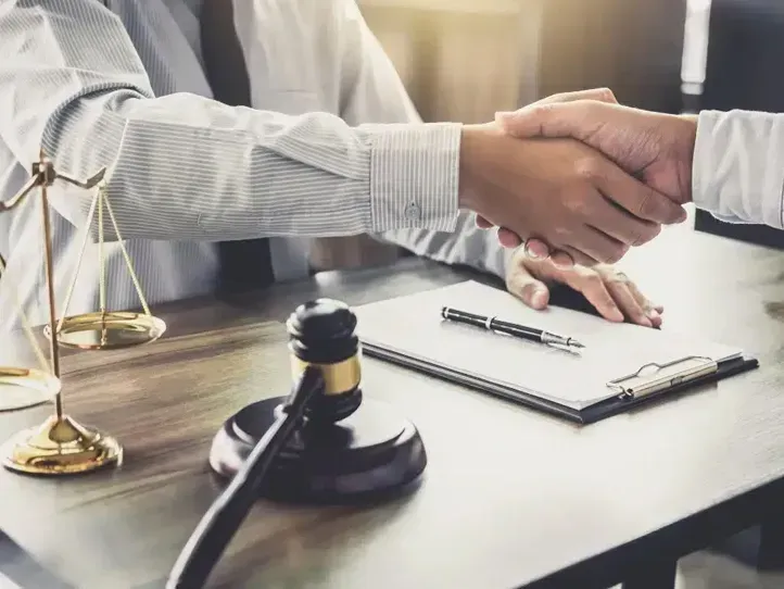 Two people in professional attire shaking hands over a desk with a wooden gavel and the scales of justice nearby.