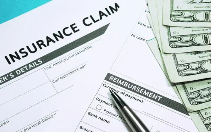 A pen rests on a printed insurance claim form next to a stack of twenty-dollar bills on a teal background.