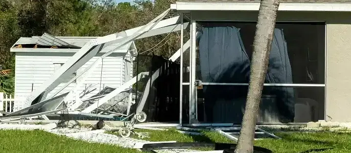 A damaged screened-in porch with broken aluminum framing and torn screens, set next to a small, damaged storage shed.