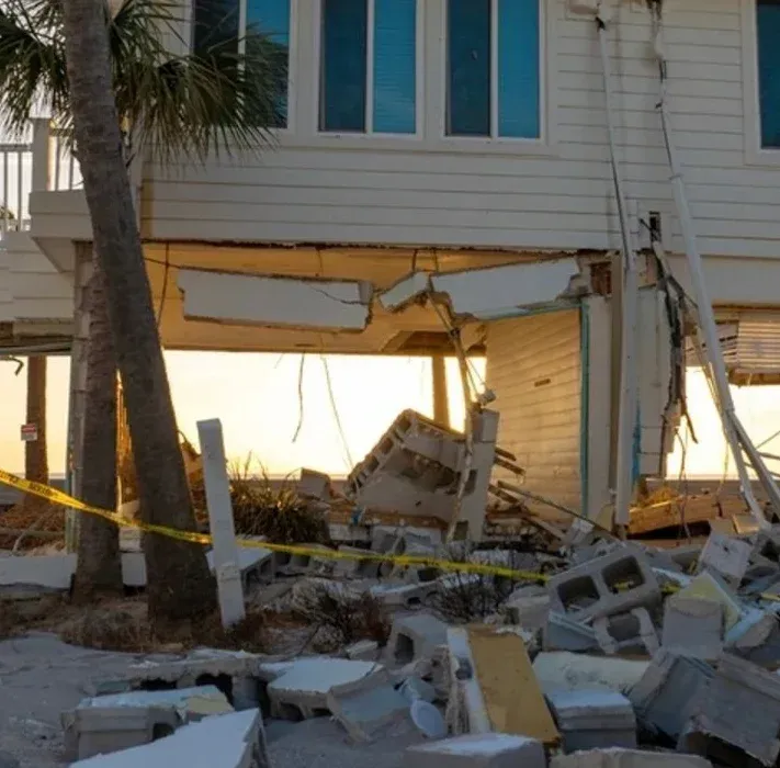 A house on pilings with its lower level destroyed by a natural disaster, leaving debris and cinder blocks in the sand.