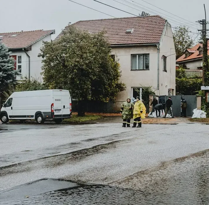 People in rain gear and workers stand near a residential house and a white van during a flood.