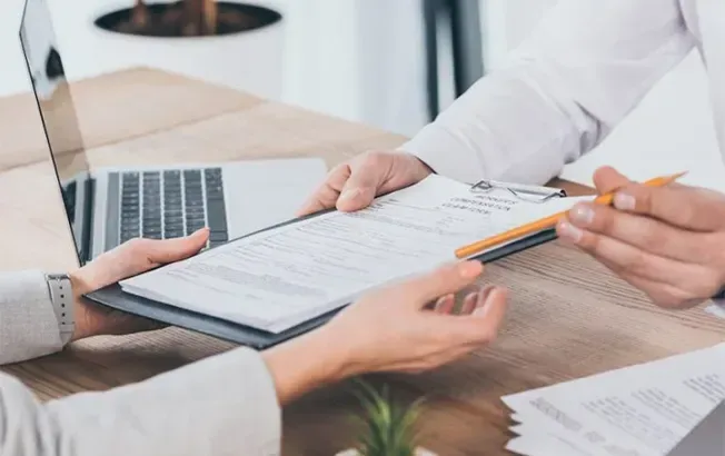 Two professionals in an office setting exchange a clipboard and pen across a wooden desk with a laptop nearby.