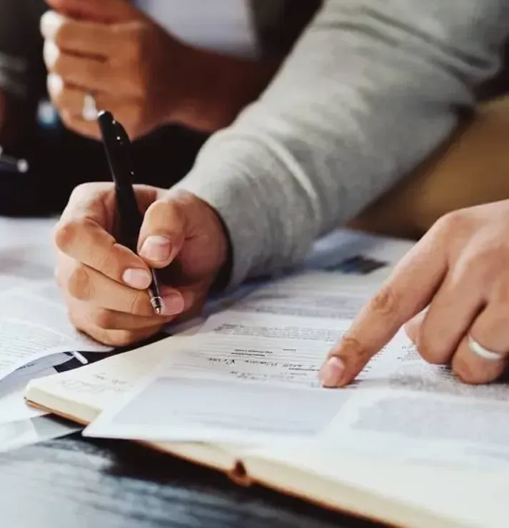 Close-up of hands writing in a document with a black pen while another hand points to the text.