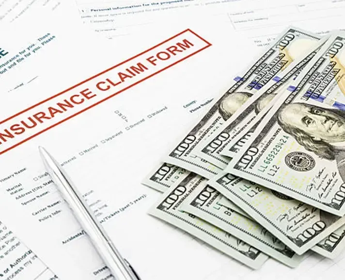 An insurance claim form sits on a table next to a silver pen and a stack of US one-hundred-dollar bills.