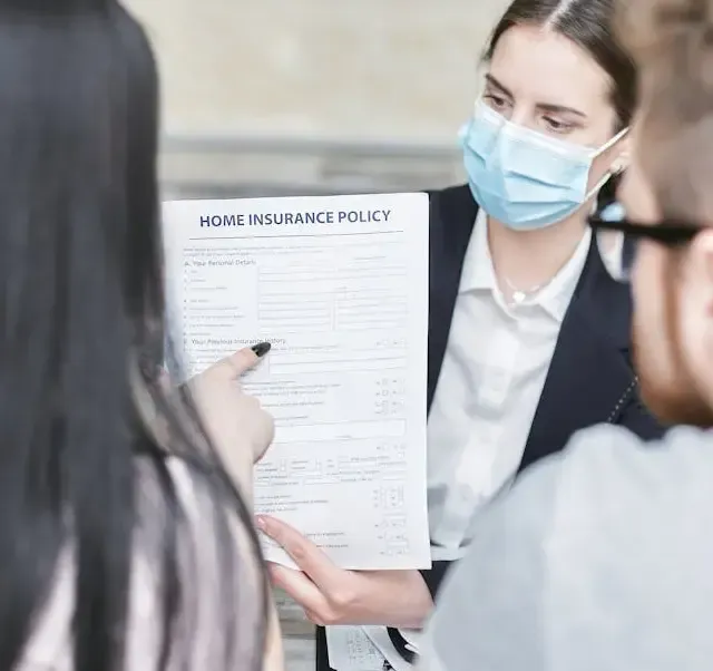 A person in a mask points to a Home Insurance Policy document while consulting with two individuals.