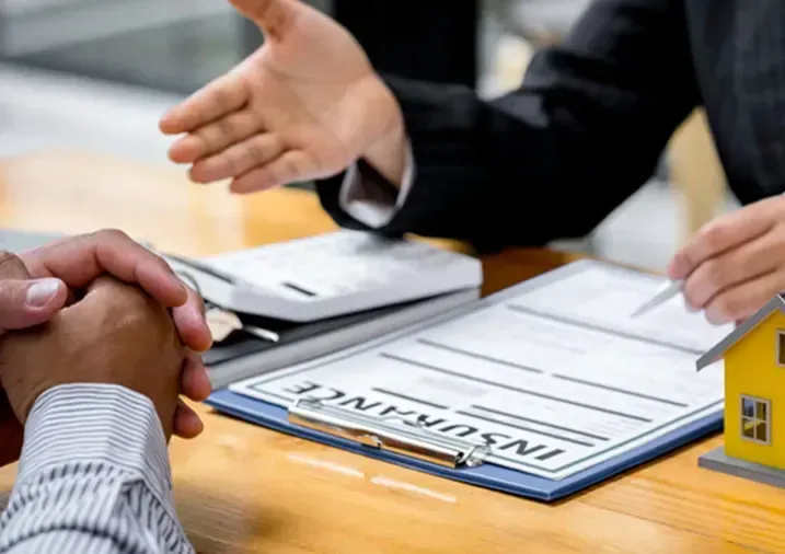 A person sitting across from an insurance agent, with a contract and a model house on a desk.