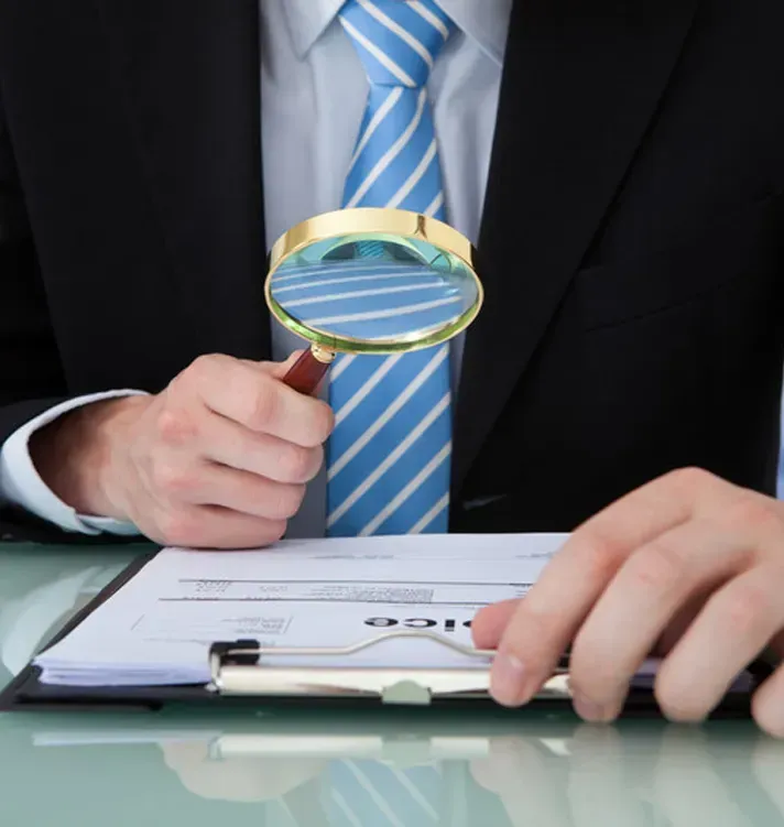 A person in a suit and striped tie holds a magnifying glass over a document on a clipboard.