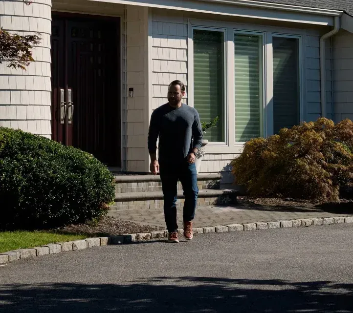 A person in a dark long-sleeved shirt and jeans walks toward the camera from the front door of a suburban house.