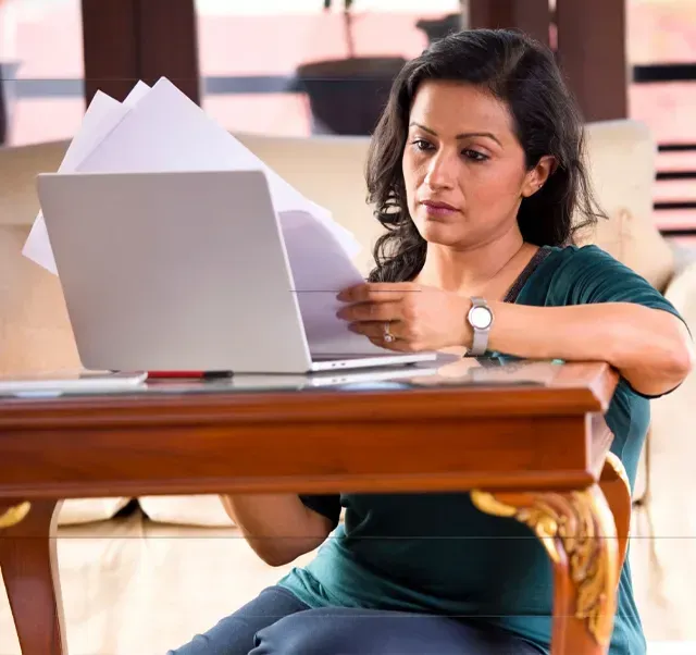 A person in a green shirt sits at a wooden table, reviewing papers while looking at an open laptop.