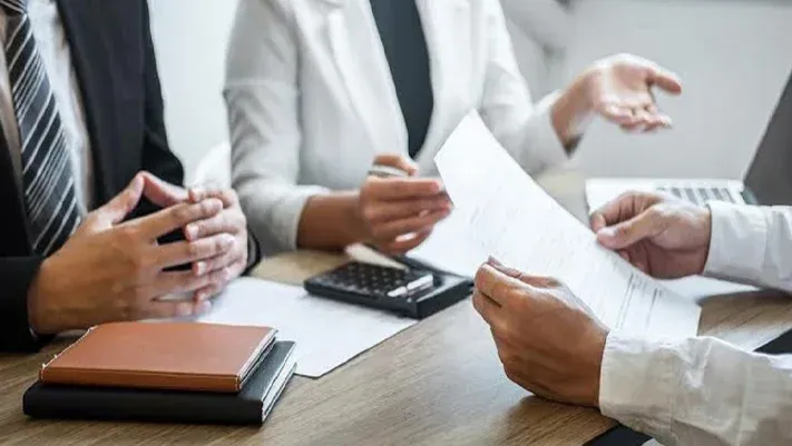 Three people in professional attire seated at a wooden desk, reviewing documents and discussing work in an office setting.
