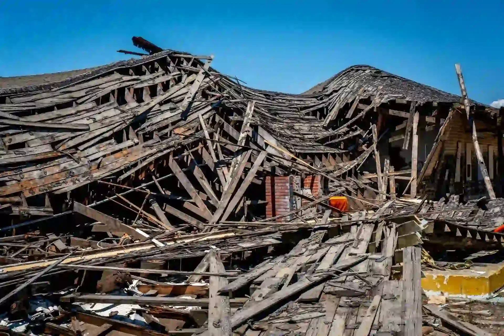 A collapsed wooden building with fragmented beams and debris under a clear blue sky.
