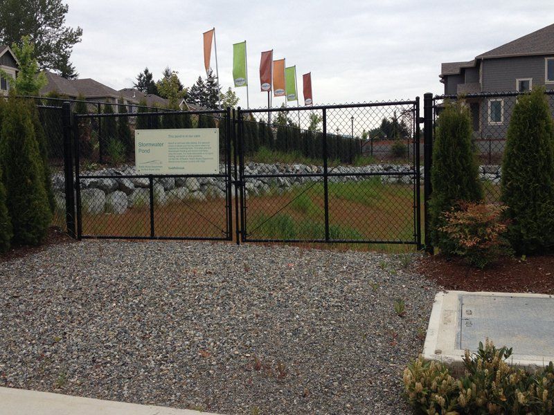 Black chain-link fence with gate, gravel pathway, and signs. Colorful flags in background.