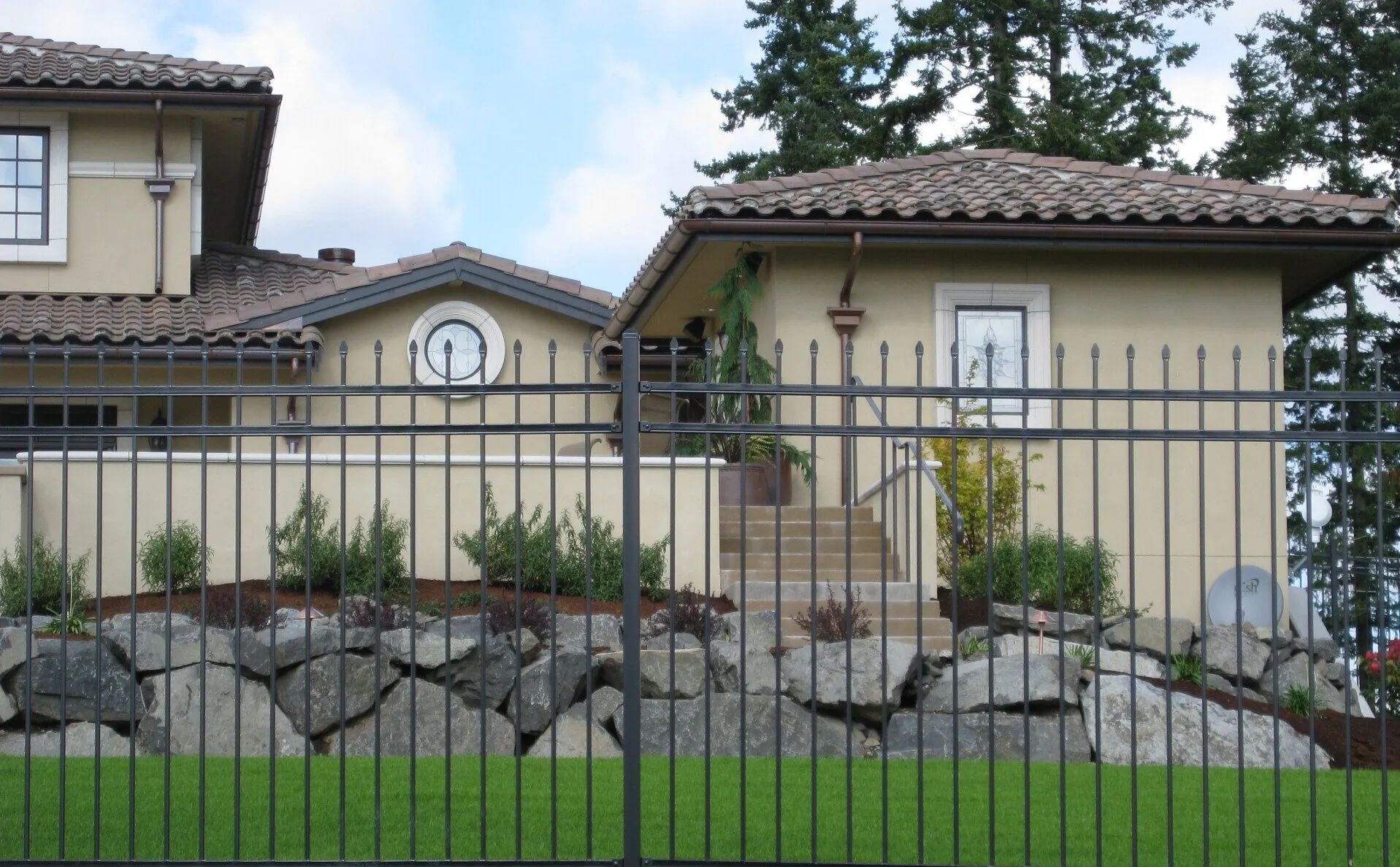 House behind a black metal fence, light tan stucco walls, tile roof, and stone retaining wall.