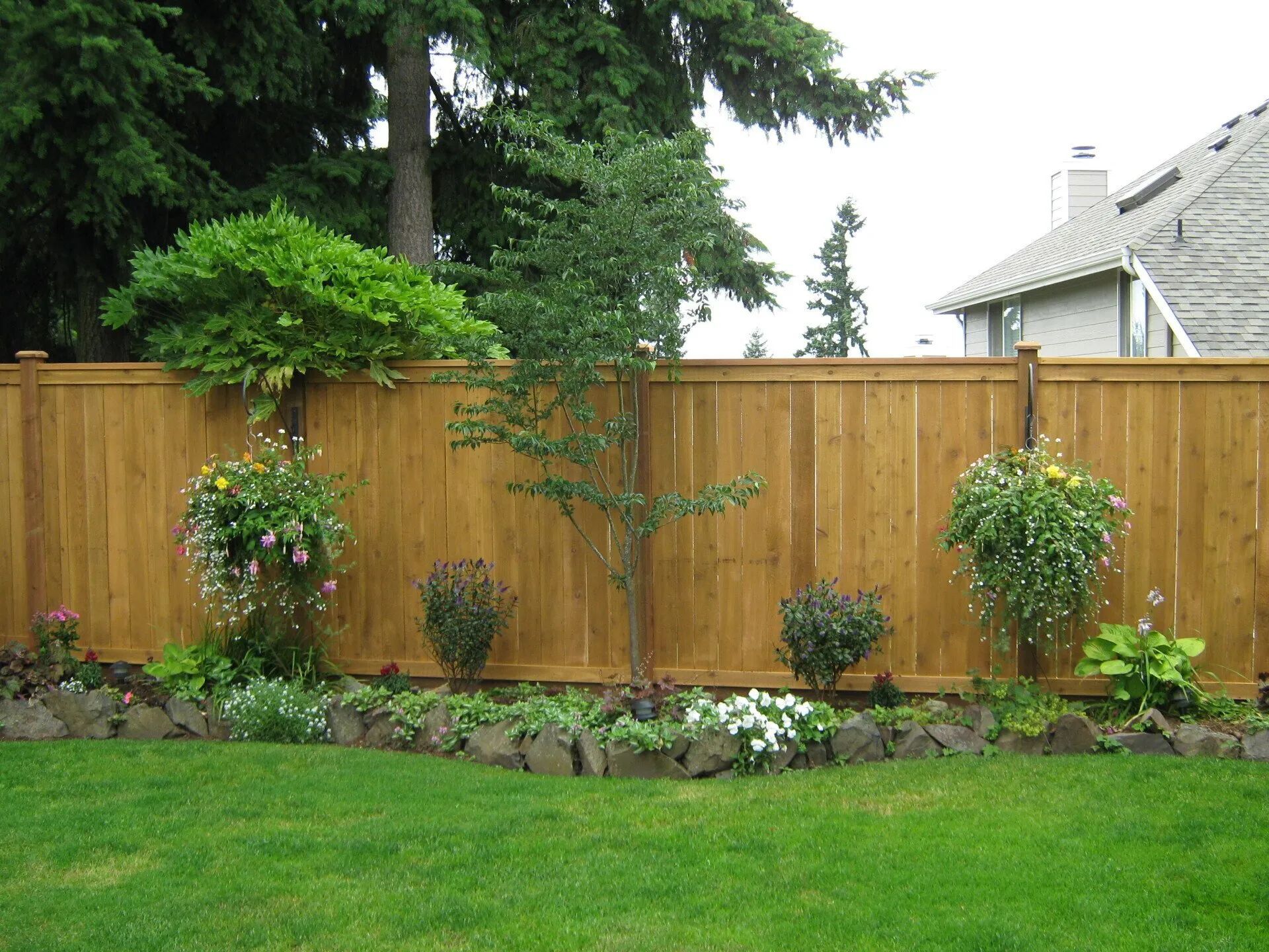 Wooden fence with a garden in front, hanging flower baskets, and green grass.