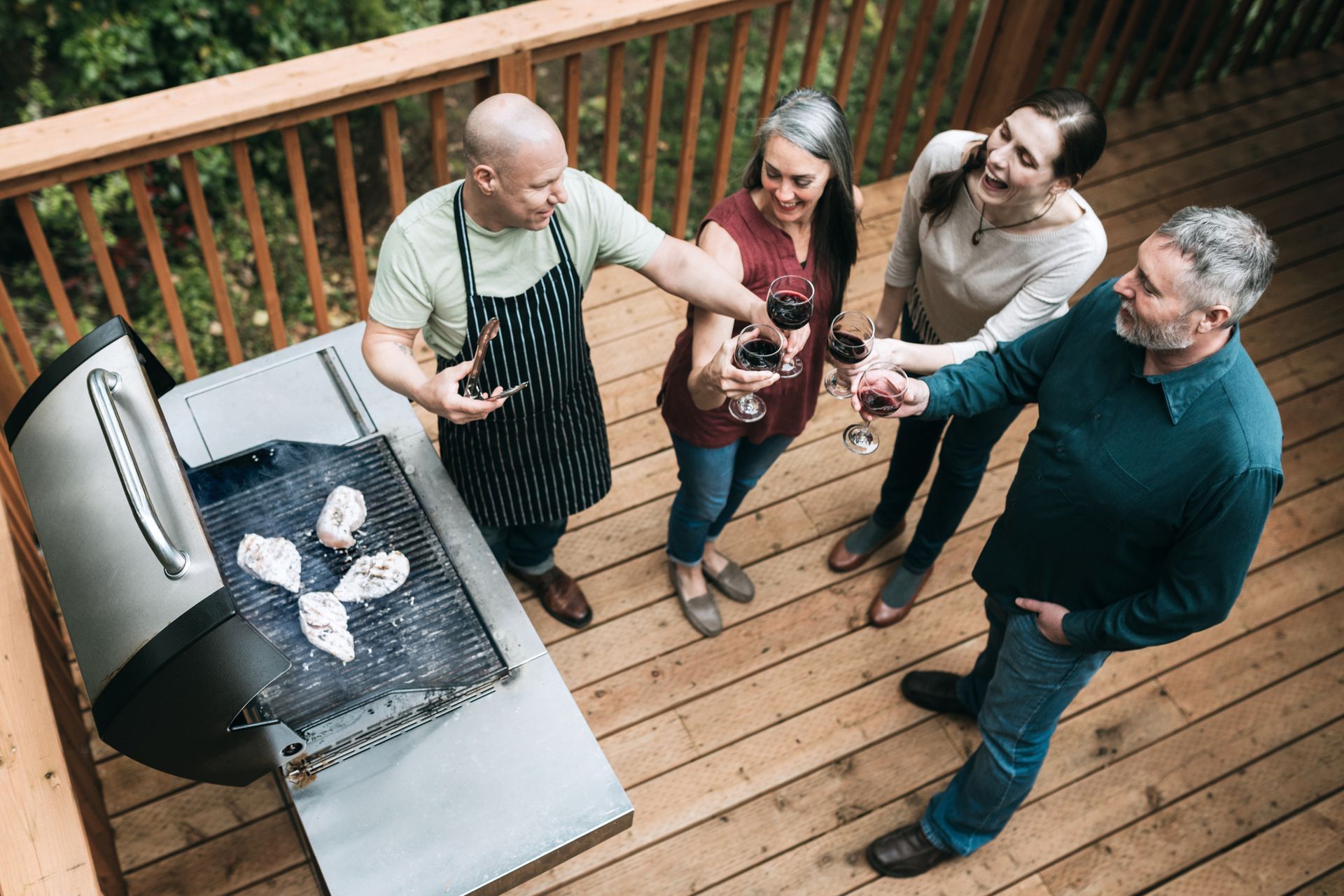 Four people on a wooden deck clink wine glasses near a grill with food.