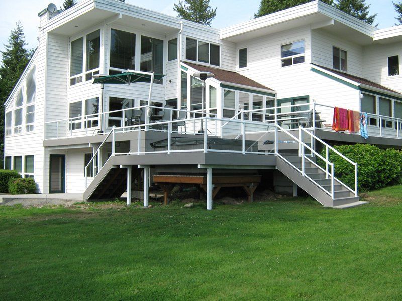 White house with large deck, glass railings, and stairs leading to a grassy yard.