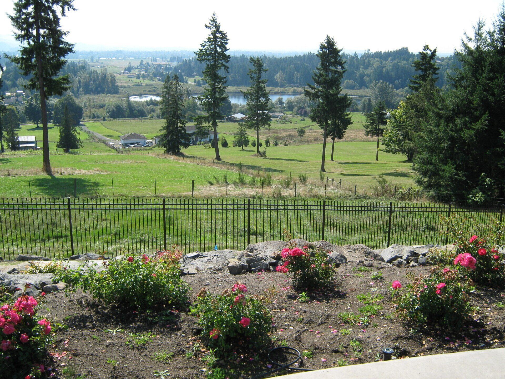 View of a green landscape with trees, a fence, and flowers in foreground, overlooking a distant body of water.