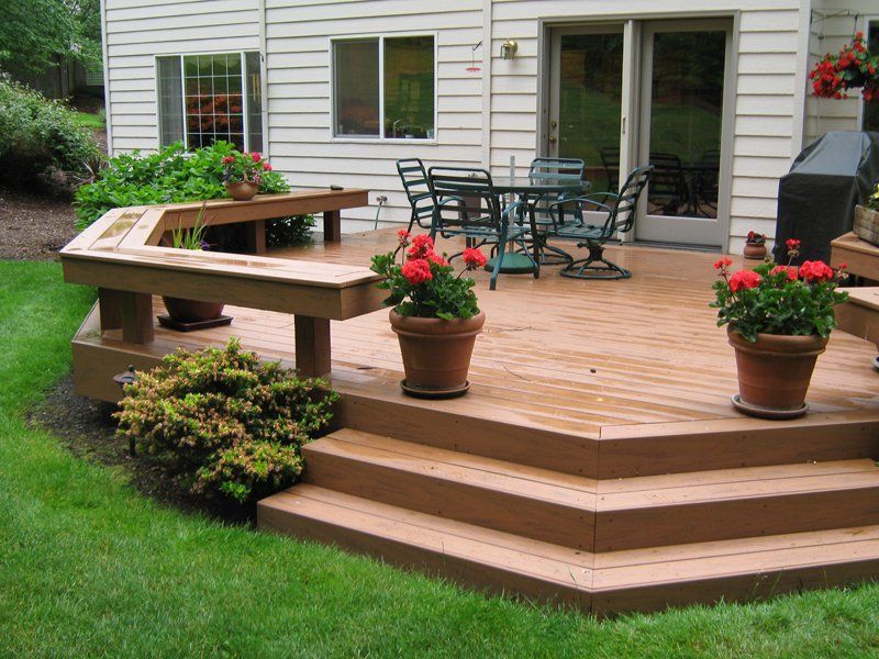 Wooden deck with steps, a built-in bench, and a dining area, surrounded by grass and potted flowers.