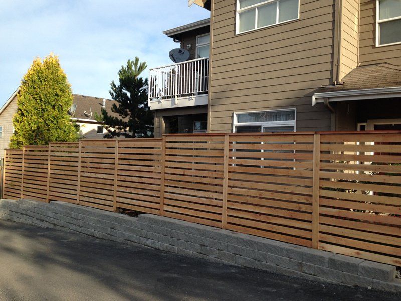 Wooden horizontal slat fence in front of a light brown building and asphalt road.