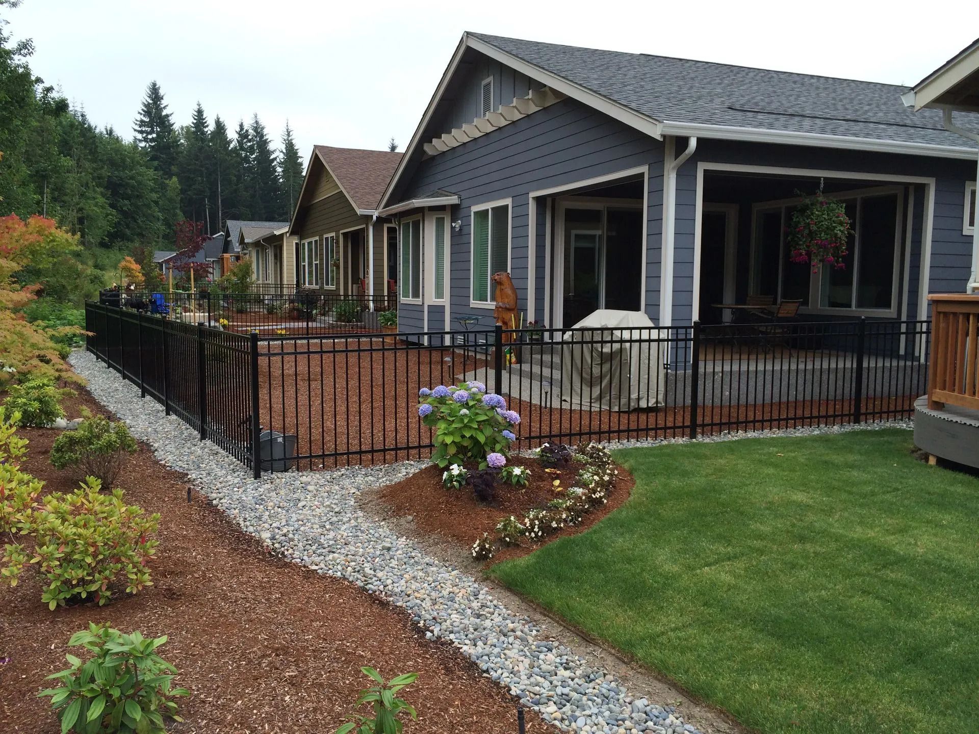 Black fence surrounds a patio and landscaped yard next to a gray house, with a forested background.