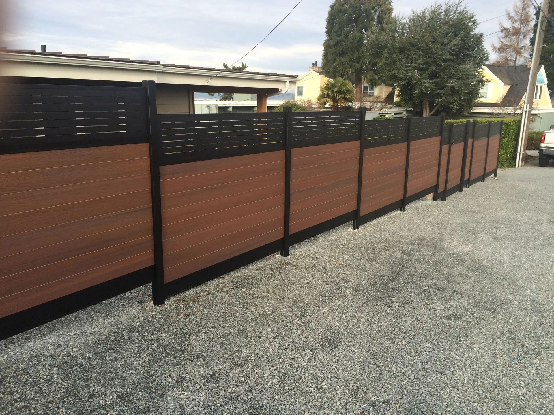 Brown and black privacy fence along a gravel driveway in front of houses.
