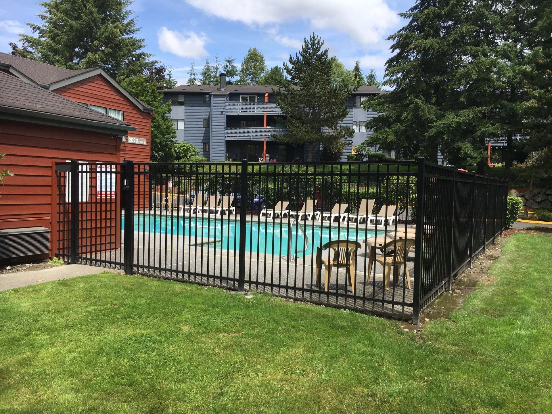 Black fenced-in swimming pool surrounded by green grass. Apartment building and red structure in the background.